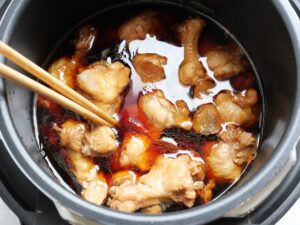 電気圧力鍋の中の鶏手羽元を、菜箸で返して甘辛の煮汁に浸している工程（圧力調理後）
